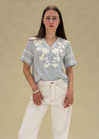 Blouse femme en coton rayé bleu et blanc à broderies florales blanches, col V boutonné, manches courtes, vue de face en pied, allure balnéaire chic Dune & Coton — Couleur: Bleu ciel
