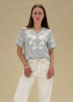 Blouse femme en coton rayé bleu et blanc à broderies florales blanches, col V boutonné, manches courtes, vue de face en pied, allure balnéaire chic Dune & Coton — Couleur: Bleu ciel